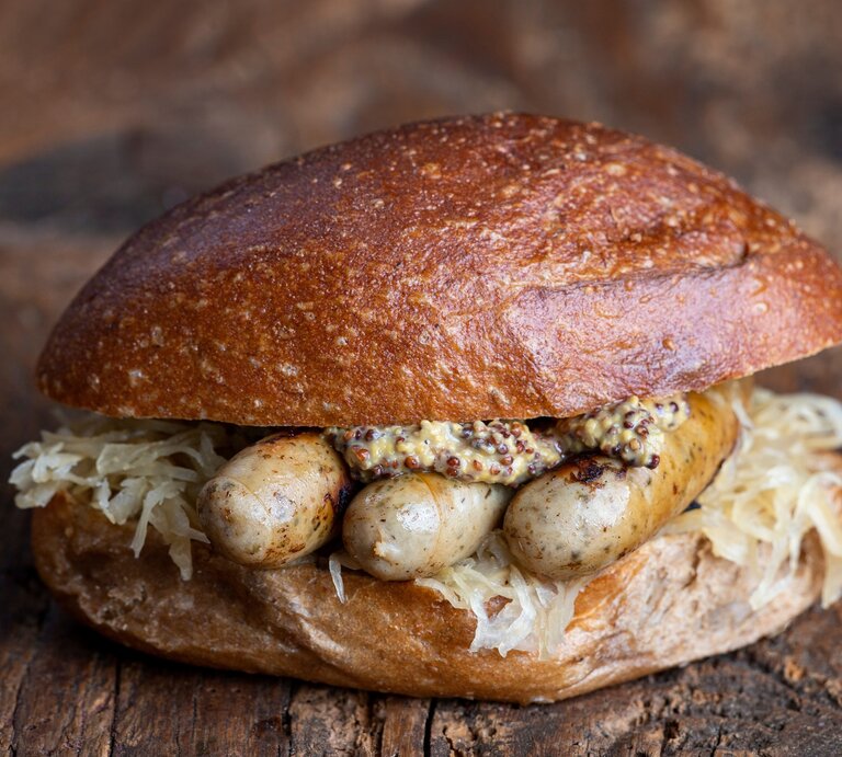 Frontal: Der fränkischer Wurstklassiker "Drei im Weggla" auf einem rustikalen Holzboden. Es handelt sich um drei Bratwürste im Brötchen mit Sauerkraut und Senf. | © Schutterstock /  Bernd Juergens