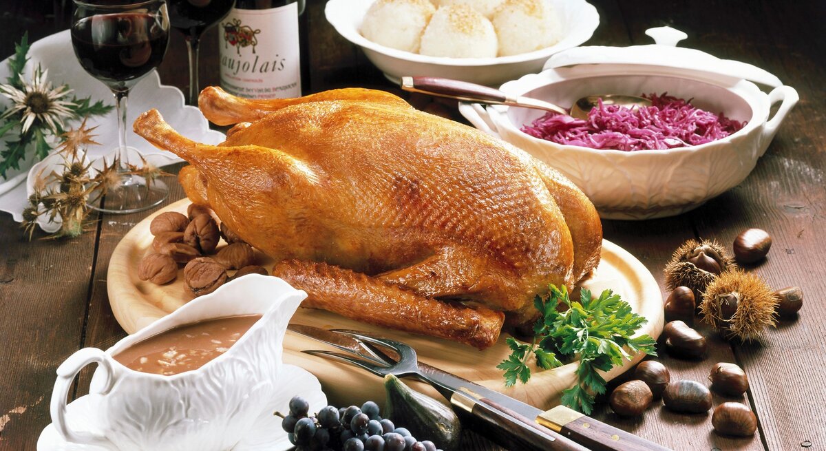 Gefüllter Gänsebraten mit Apfel und Marone auf Festtafel mit Beilagen. Im Hintergrund eine Flasche Rotwein und zwei Gläser und Dekoration. Draufsicht.