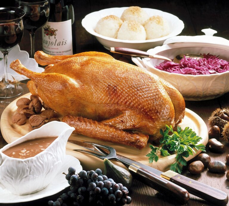 Gefüllter Gänsebraten mit Apfel und Marone auf Festtafel mit Beilagen. Im Hintergrund eine Flasche Rotwein und zwei Gläser und Dekoration. Draufsicht. | © Getty Images / StockFood