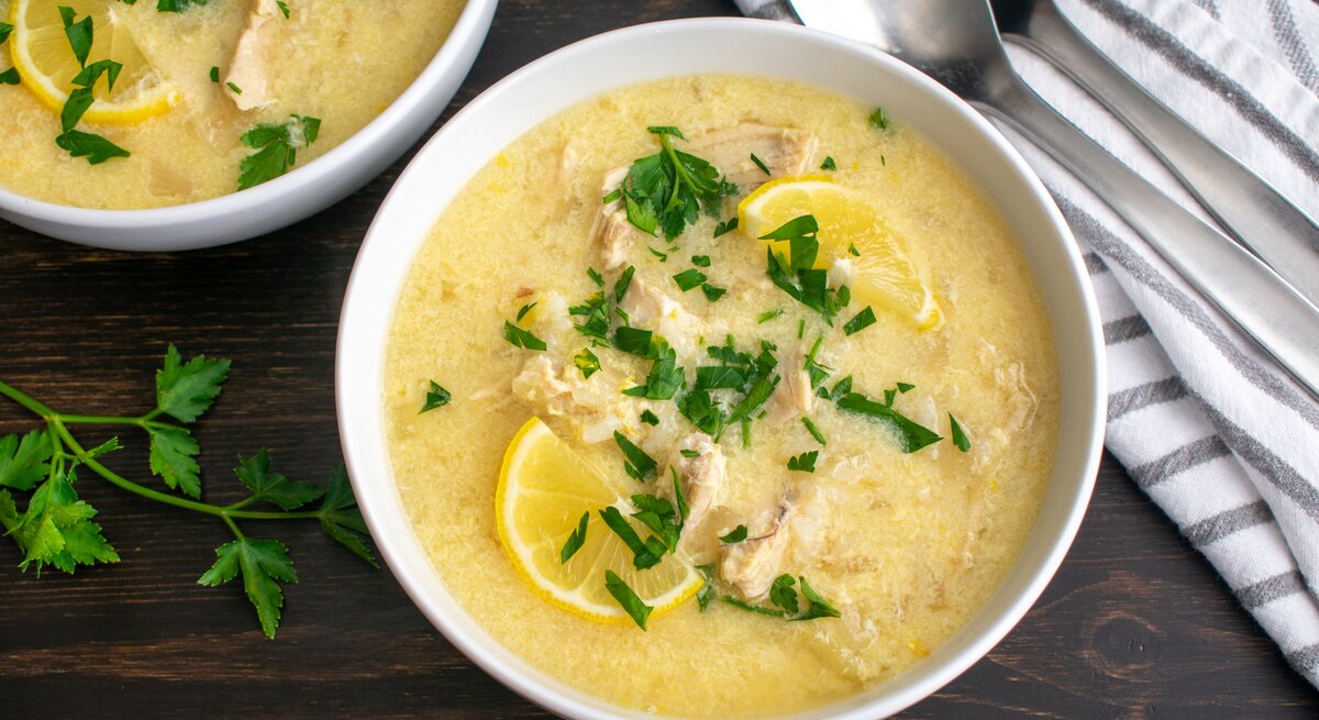 Draufsicht: Zwei Schüsseln mit der mediterranen Hühnersuppe Avgolemono stehen auf einem dunklen Holztisch. Die Suppe ist mit Petersilie und Zitronenscheiben garniert. Daneben liegen zwei Löffel und ein gestreiftes Küchenhandtuch.