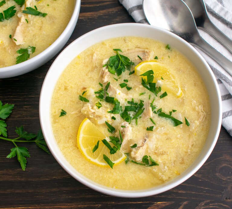 Draufsicht: Zwei Schüsseln mit der mediterranen Hühnersuppe Avgolemono stehen auf einem dunklen Holztisch. Die Suppe ist mit Petersilie und Zitronenscheiben garniert. Daneben liegen zwei Löffel und ein gestreiftes Küchenhandtuch. | © Shutterstock / Candice Bell