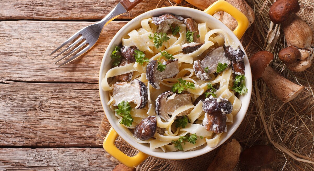 Draufsicht: Auf einem Holzboden steht ein Topf mit Tagliatelle mit Steinpilzen. Drum herum liegen eine Gabel und frische Steinpilze.