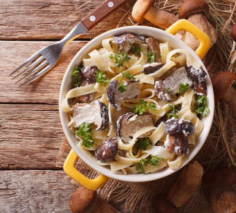 Draufsicht: Auf einem Holzboden steht ein Topf mit Tagliatelle mit Steinpilzen. Drum herum liegen eine Gabel und frische Steinpilze. | © Shutterstock / AS Foodstudio