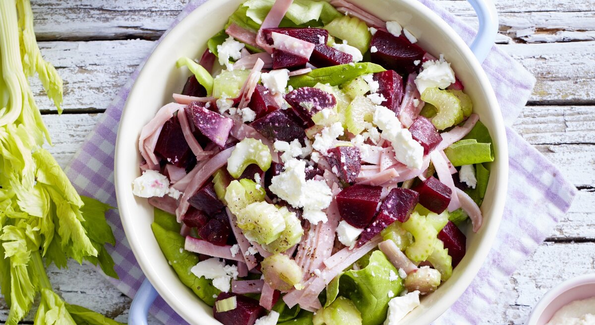 Salat mit Rote Bete und Schinken, getoppt mit frischem Feta und Sellerie, serviert in einer großen weißen Schale mit Henkeln auf einem Holztisch. 