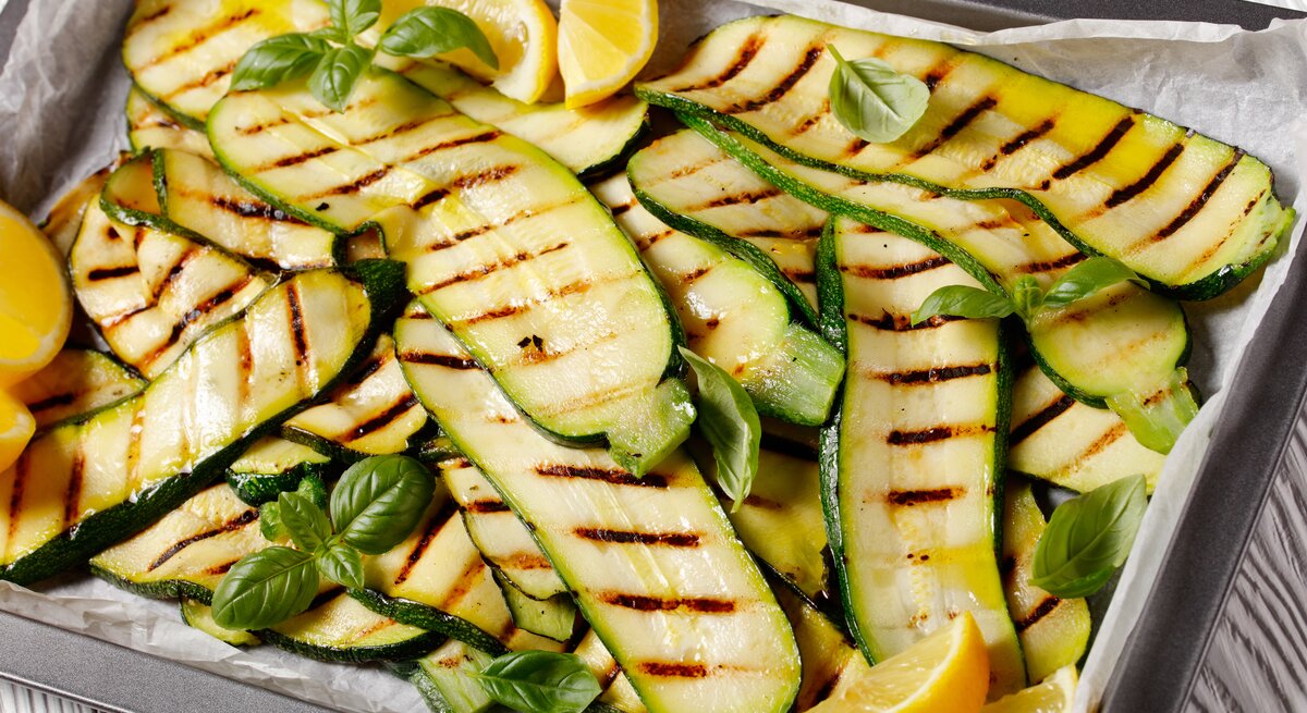 Auf einem mit Backpapier ausgekleideten Backblech liegen Zucchini, die gegrillt worden sind. Auf und zwischen ihnen liegen Zirtonenschnitzer und Basilikumblätter.