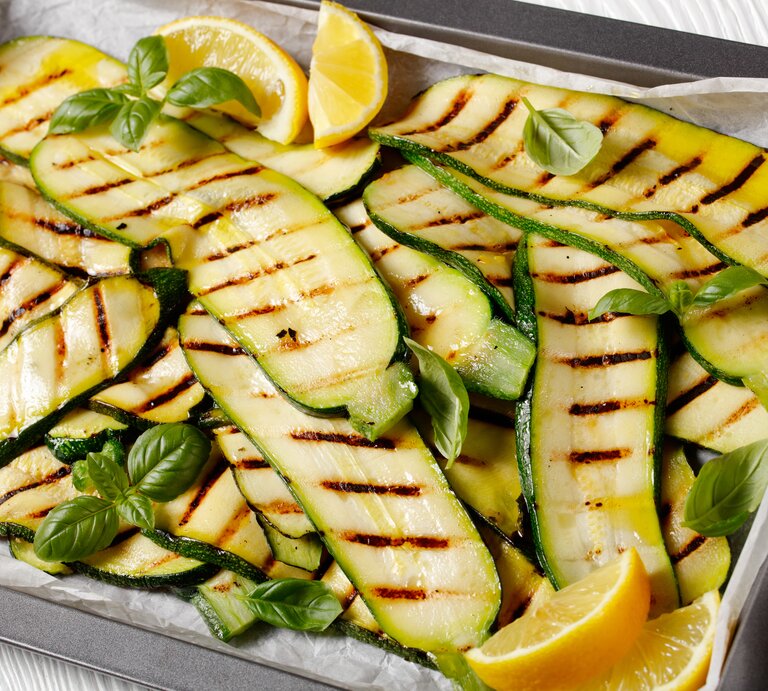 Auf einem mit Backpapier ausgekleideten Backblech liegen Zucchini, die gegrillt worden sind. Auf und zwischen ihnen liegen Zirtonenschnitzer und Basilikumblätter. | © Shutterstock / from my point of view