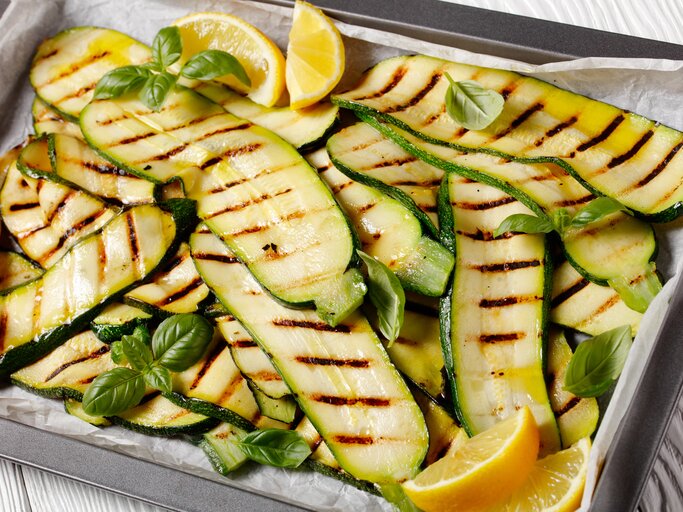 Auf einem mit Backpapier ausgekleideten Backblech liegen Zucchini, die gegrillt worden sind. Auf und zwischen ihnen liegen Zirtonenschnitzer und Basilikumblätter. | © Shutterstock / from my point of view