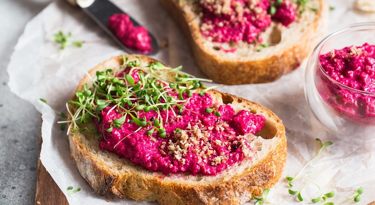 Ein paar Brotscheiben mit Pink Hummus, gehackten Nüssen und Kresse auf einem Holzbrett.