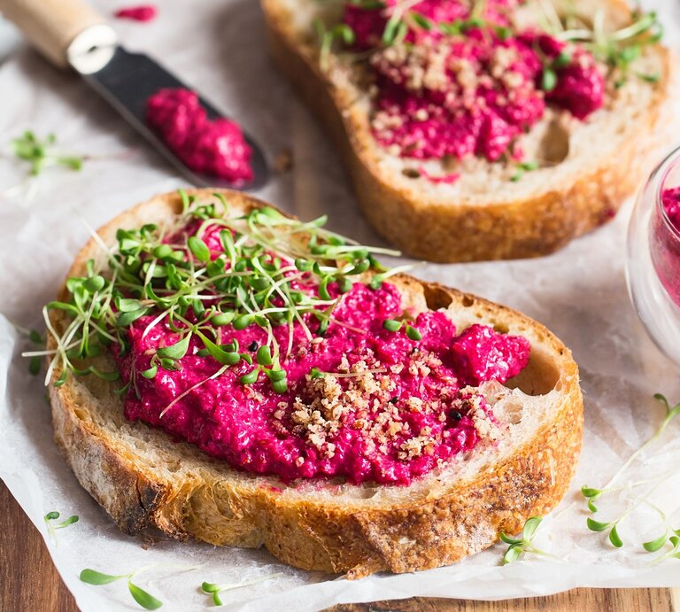 Ein paar Brotscheiben mit Pink Hummus, gehackten Nüssen und Kresse auf einem Holzbrett. | © Adobe Stock/ nata_zhekova