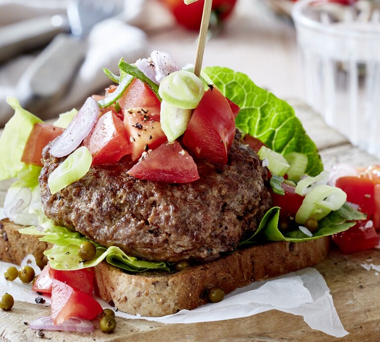 Leichter Beef-Burger mit grünem Pfeffer auf Holzunterlage, im Hintergrund Geschirr. | © EAT CLUB / Isabell Triemer