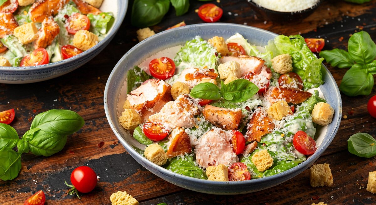 Zwei Teller mit dem Caesar Salat auf einem dunklen Holztisch. Drum herum Salat, Kräuter und Kirschtomaten.