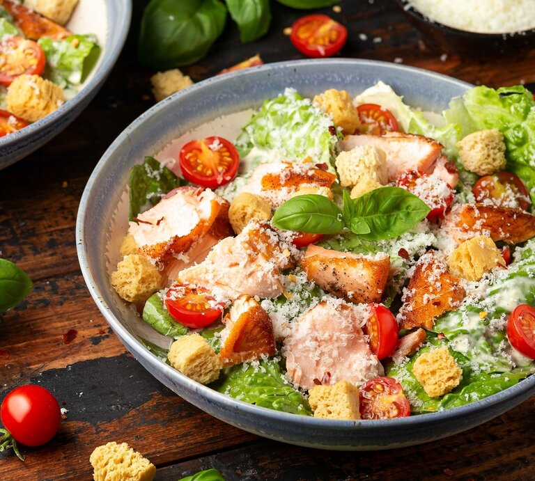 Zwei Teller mit dem Caesar Salat auf einem dunklen Holztisch. Drum herum Salat, Kräuter und Kirschtomaten. | © Shutterstock/ DronG