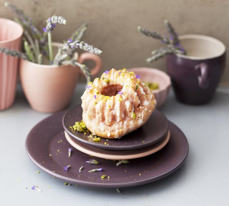 Ein Himbeer-Lavendel-Minigugelhupf auf Tellerstapel mit Geschirr und Lavendel im Hintergrund. Weißer Untergrund und beige Wand. | © © StockFood / Gräfe & Unzer Verlag / Grossmann.Schuerle