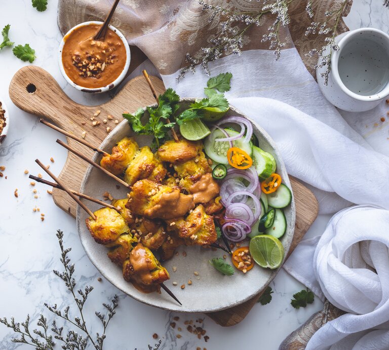 Ein Teller mit fünf Erdnuss-Satay-Spießen mit Salat, garniert mit Erdnussstücken, Koriander und zusätzlicher Erdnusssoße, die in einer kleinen Schüssel dazugereicht wird. Der Teller steht auf einem Holzschneidebrett, neben einem hellen Tuch vor einem weißen Hintergrund, von oben.  | © Getty Images/asab974