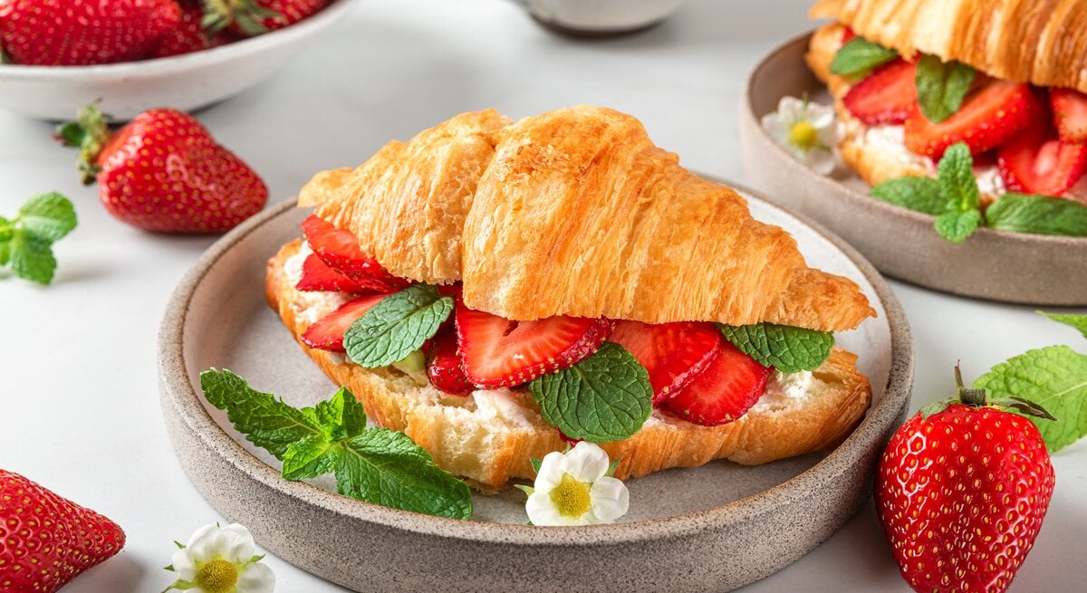 Frontalsicht: Ein Erdbeer-Croissant mit frischer Minze liegt in einer runden Tonschüssel, die auf einem weißen Untergrund liegt. Drum herum liegen viele frische Erdbeeren und weiße Erdbeerblüten.