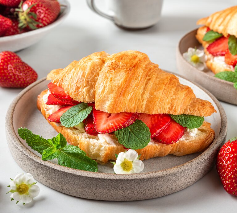 Frontalsicht: Ein Erdbeer-Croissant mit frischer Minze liegt in einer runden Tonschüssel, die auf einem weißen Untergrund liegt. Drum herum liegen viele frische Erdbeeren und weiße Erdbeerblüten. | © Shutterstock / artem evdokimov