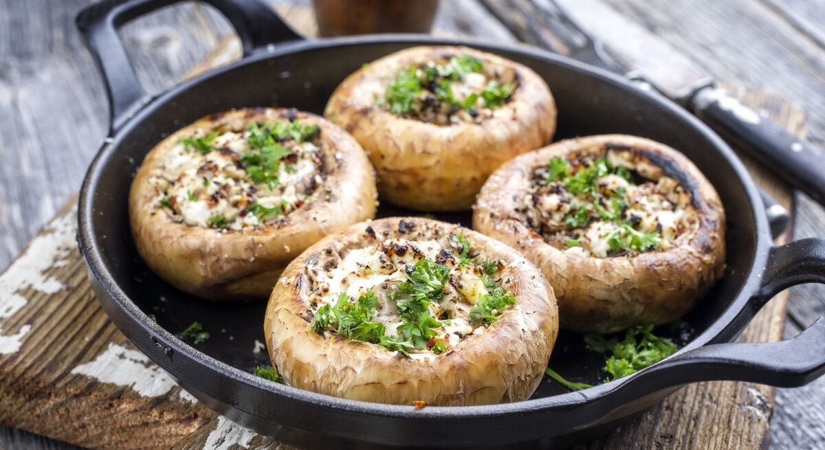 Eine gusseiserne Pfanne mit vier gefüllten Champignons auf einem dunklen Holztisch von schräg oben fotografiert.