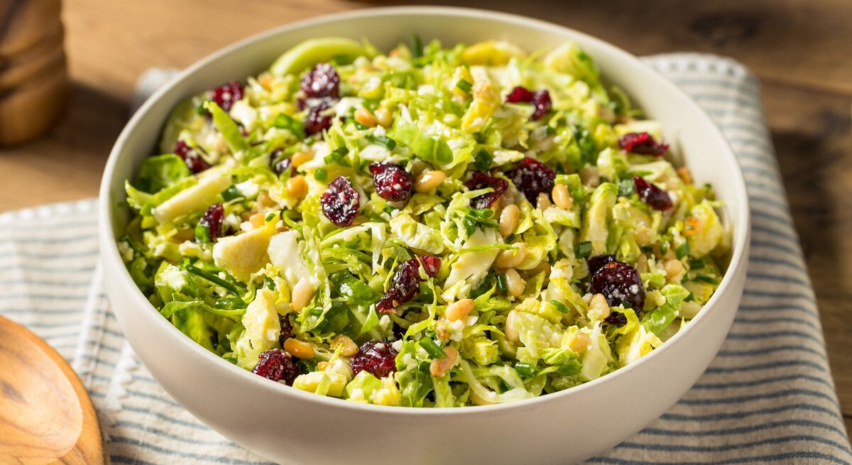 Eine weiße Schale mit dem Rosenkohl-Salat mit Cranberries und Pinienkernen auf einem blau-weiß gestreiftem Tuch und einem Holztisch von oben fotografiert.