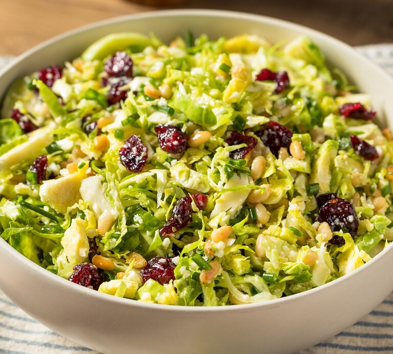 Eine weiße Schale mit dem Rosenkohl-Salat mit Cranberries und Pinienkernen auf einem blau-weiß gestreiftem Tuch und einem Holztisch von oben fotografiert. | © Shutterstock/ Brent Hofacker  