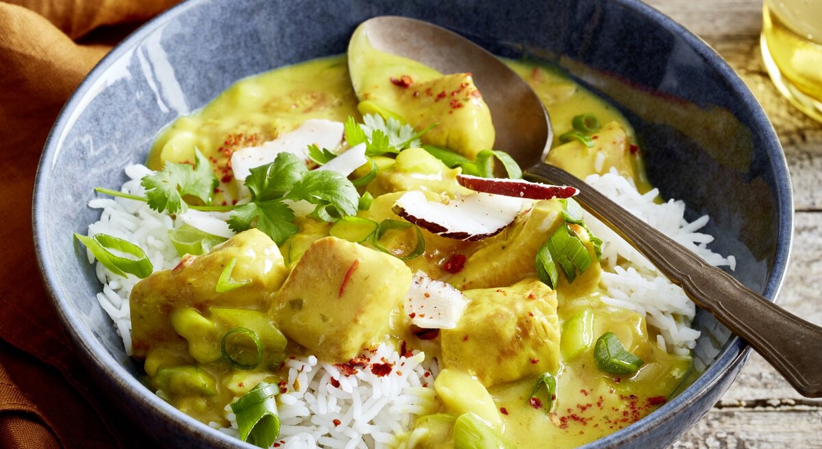 Eine Schüssel mit Basmati-Reis und cremigem Curry mit Hähnchenstücken, garniert mit frischen Frühlingszwiebeln, Koriander, Chiliflocken und Kokosraspeln. Eine Gabel liegt am Schüsselrand, im Hintergrund ist ein Getränk zu sehen.