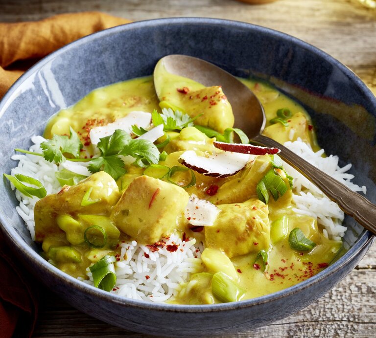 Eine Schüssel mit Basmati-Reis und cremigem Curry mit Hähnchenstücken, garniert mit frischen Frühlingszwiebeln, Koriander, Chiliflocken und Kokosraspeln. Eine Gabel liegt am Schüsselrand, im Hintergrund ist ein Getränk zu sehen. | © EAT CLUB / Isabell Triemer  