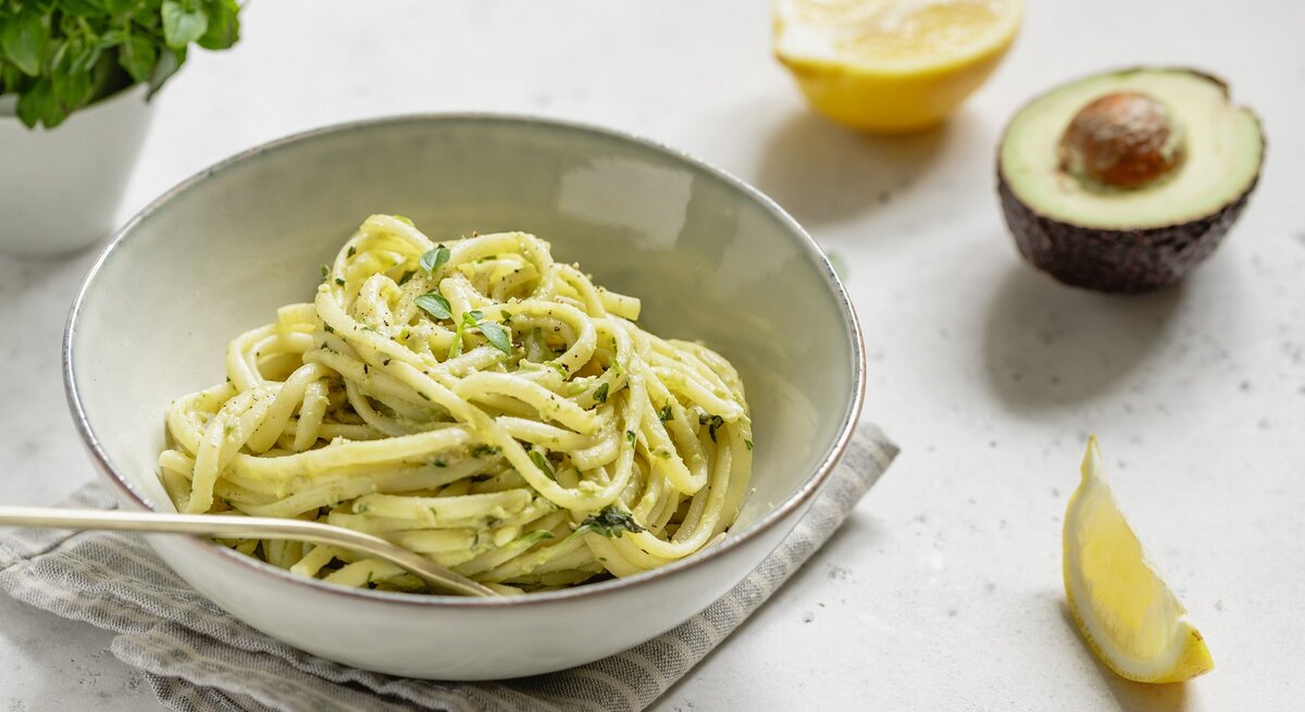 Ein weißer Teller mit Avocado-Lemon-Pasta auf einem weißen Tisch neben frischer Avocado und Zitrone.