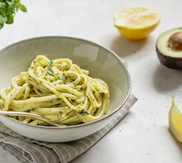 Ein weißer Teller mit Avocado-Lemon-Pasta auf einem weißen Tisch neben frischer Avocado und Zitrone. | © AdobeStock/Edalin
