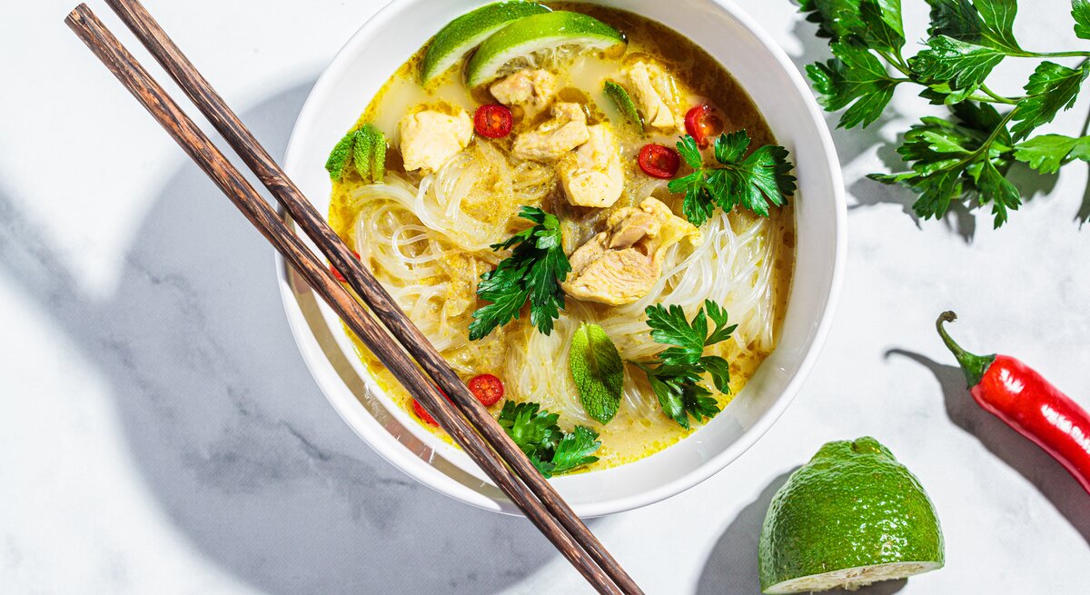 Eine weiße Schüssel mit Thai-Currysuppe mit Nudeln auf einem weißen Tisch.