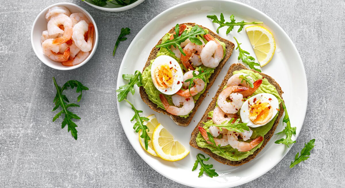 Ein weißer Teller mit den belegten Brotscheiben auf einem grauen Steintisch mit einem Schälchen mit Garnelen und einem mit Rucola direkt von oben fotografiert.