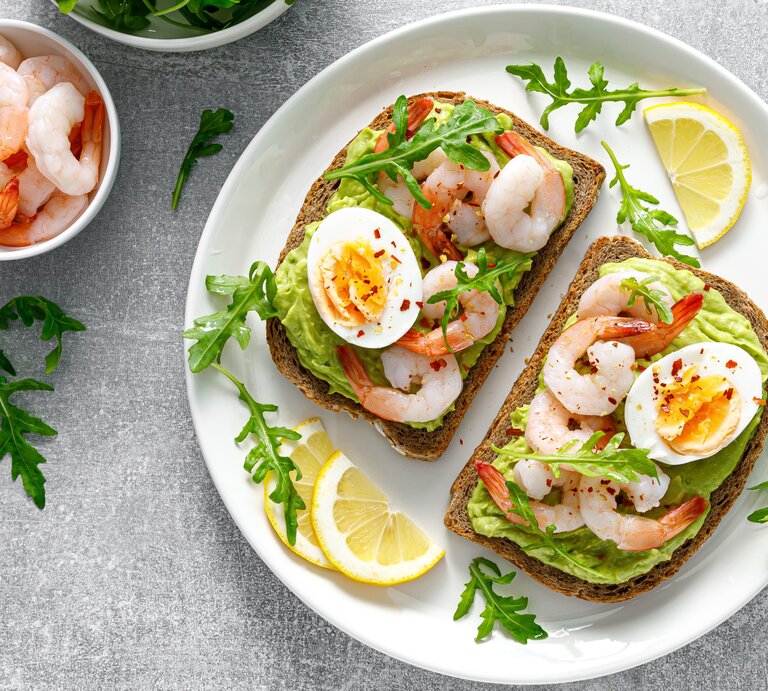 Ein weißer Teller mit den belegten Brotscheiben auf einem grauen Steintisch mit einem Schälchen mit Garnelen und einem mit Rucola direkt von oben fotografiert. | © Adobe Stock/ Sea Wave