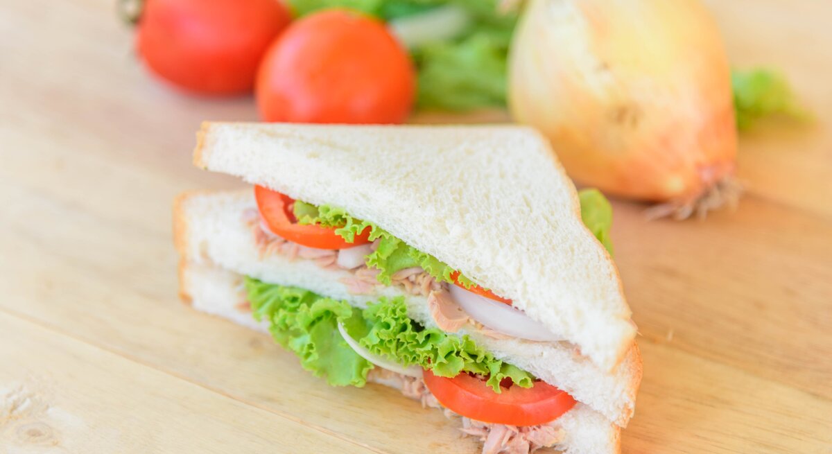 Draufsicht, schräg: Ein Thunfisch-Tramezzini mit Tomate auf einem hellem Untergrund aus Holz.
