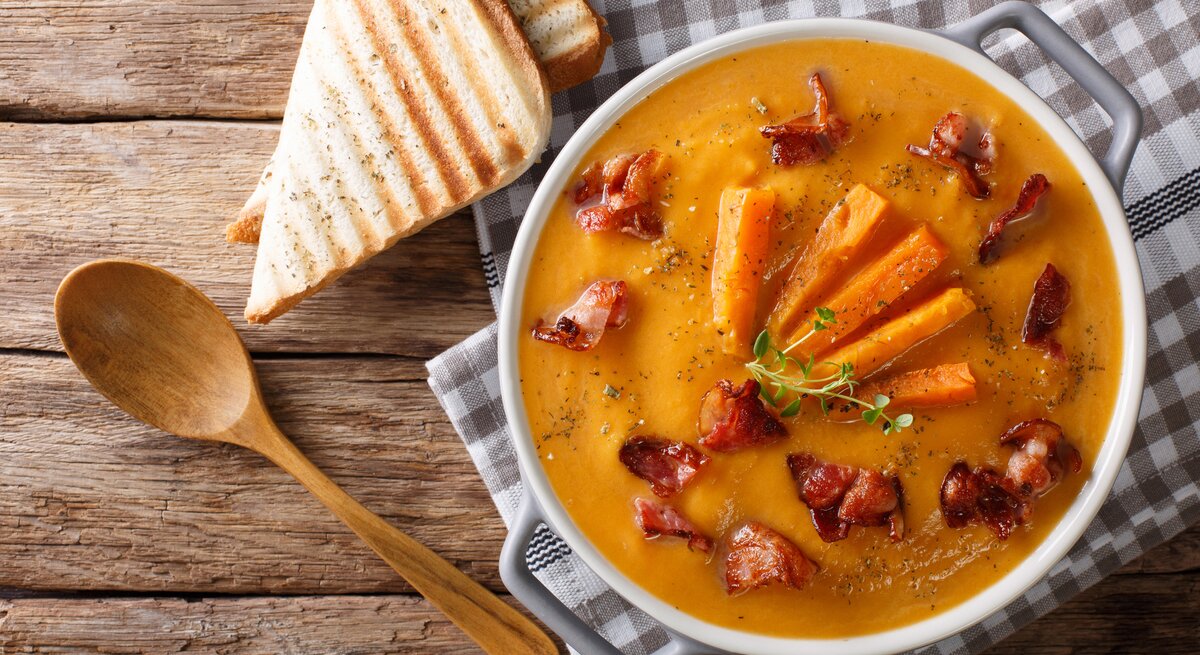 In einer großen Schüssel ist eine Süßkartoffel-Rahmsuppe. Die Suppenschüssel steht auf einem karierten Handtuch, dass auf einem Holztisch liegt. Daneben liegen zwei Scheiben Toast und ein Holzlöffel.
