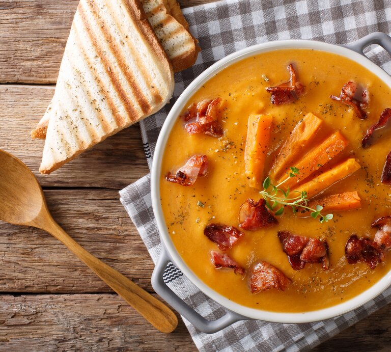 In einer großen Schüssel ist eine Süßkartoffel-Rahmsuppe. Die Suppenschüssel steht auf einem karierten Handtuch, dass auf einem Holztisch liegt. Daneben liegen zwei Scheiben Toast und ein Holzlöffel. | © Shutterstock / FomaA