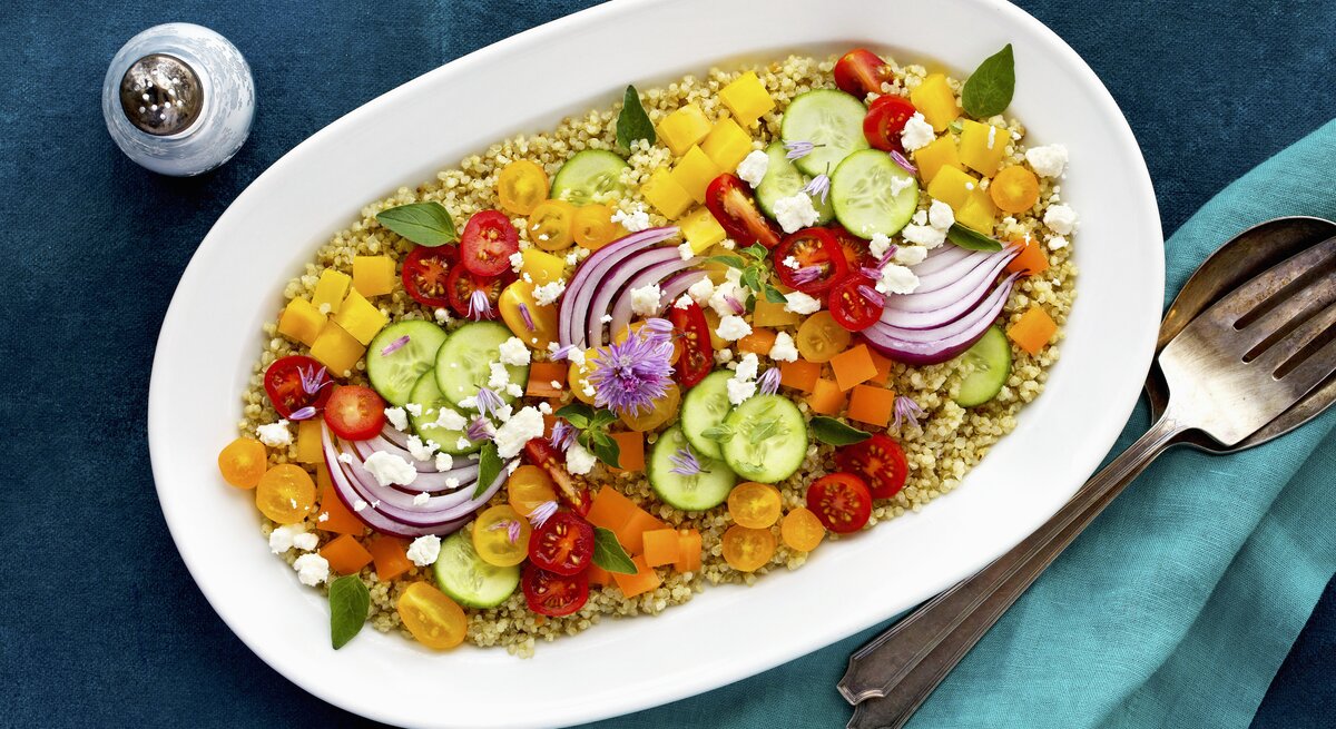 Bunter Salat aus Tomate, Paprika und Gurke auf Quinoa mit Fetakäse angerichtet auf einem weißen ovalen Porzellanteller, der auf blauer Tischedecke steht, neben dem Teller liegt Silberbesteck und steht ein Salzstreuer