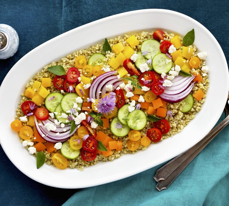 Bunter Salat aus Tomate, Paprika und Gurke auf Quinoa mit Fetakäse angerichtet auf einem weißen ovalen Porzellanteller, der auf blauer Tischedecke steht, neben dem Teller liegt Silberbesteck und steht ein Salzstreuer | © StockFood / The Picture Pantry