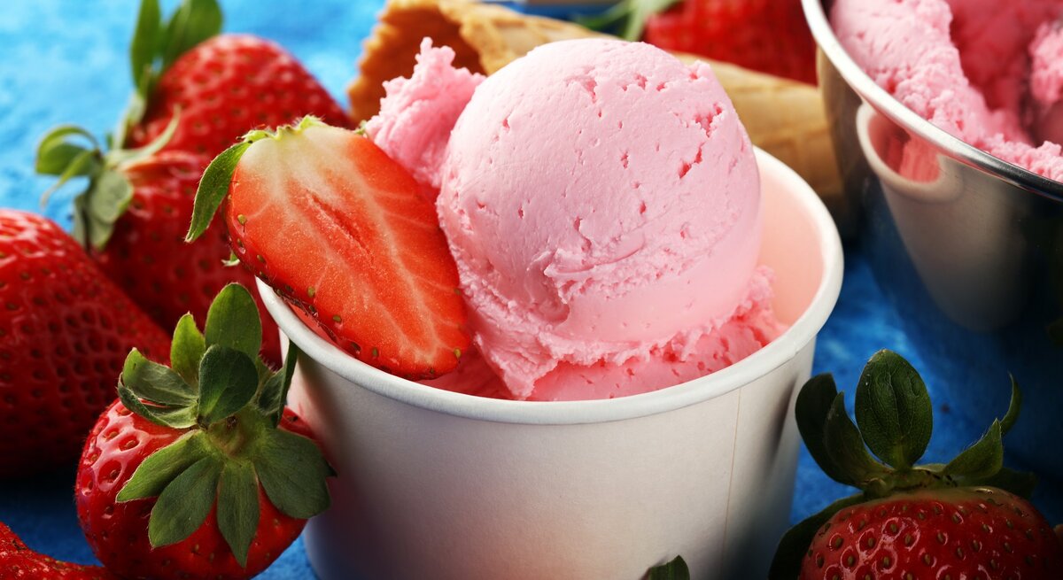 Der Erdbeer-Eisbecher zwischen vielen Erdbeeren und einer Waffel auf einem blauen Tisch.