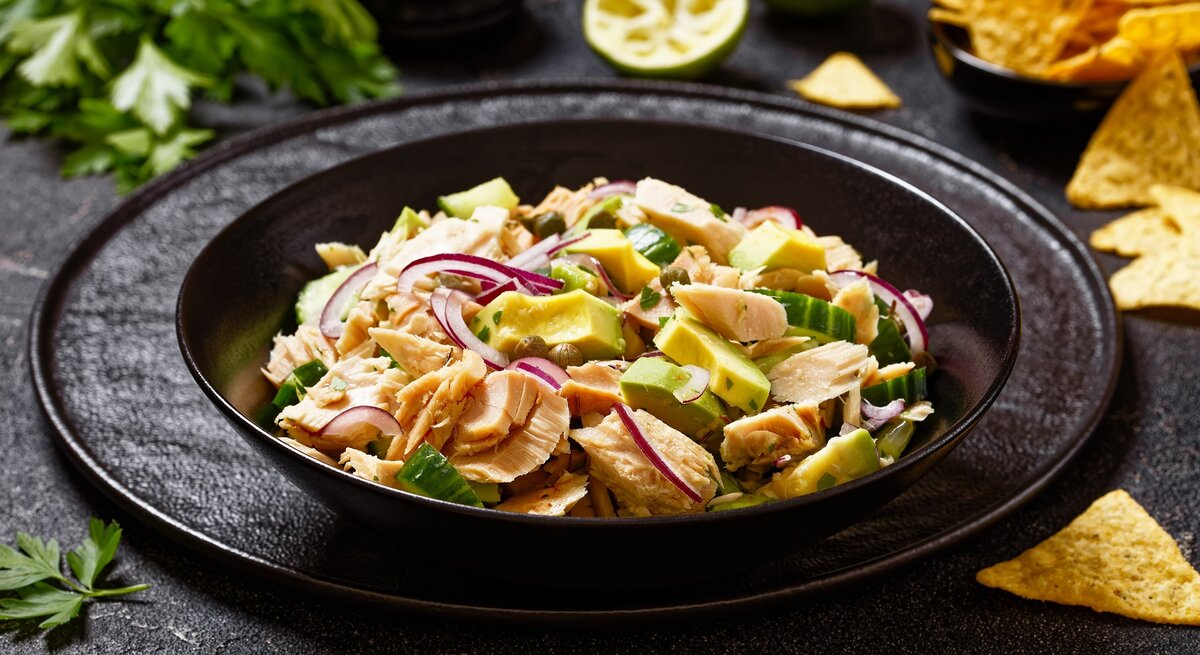 Ein schwarzer Teller mit dem Thunfischsalat, Avocado und Kapern auf einen schwarzen Tisch mit Limette, Nachos und  Petersilie.
