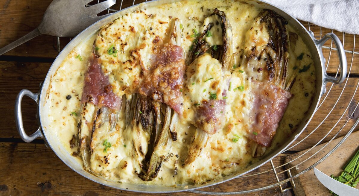 Keto Gratin mit Chicorée in Metall-Auflaufform auf Gitter, Holzuntergrund und Servierbesteck daneben. 
