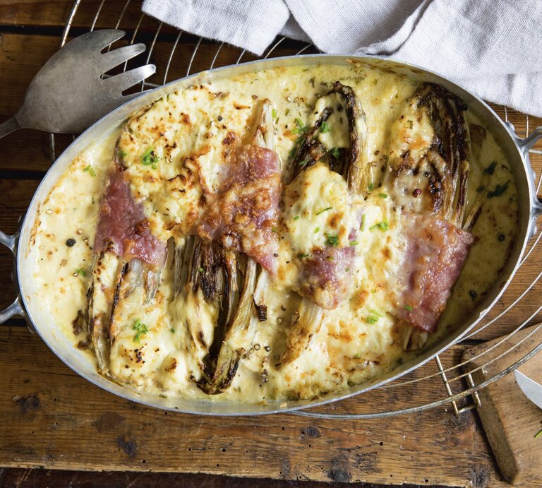 Keto Gratin mit Chicorée in Metall-Auflaufform auf Gitter, Holzuntergrund und Servierbesteck daneben.  | © StockFood / ZS Verlag / Claud Timmann
