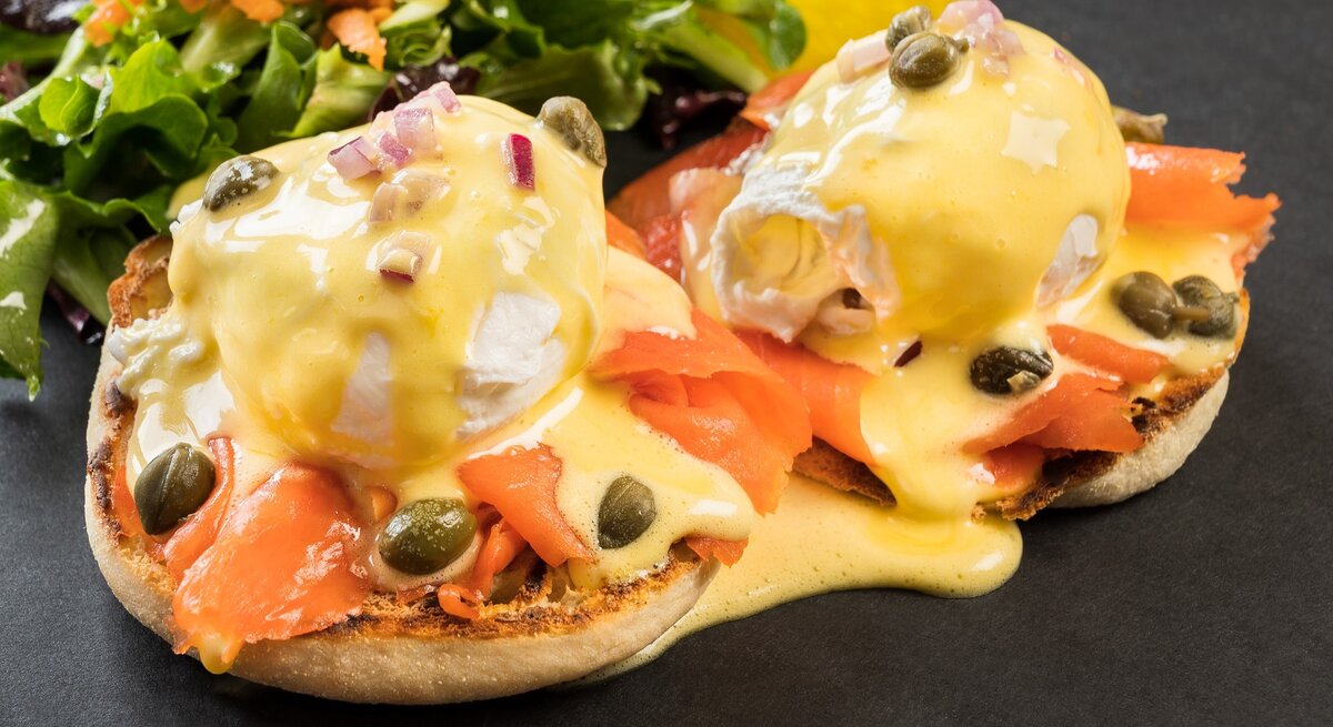 Die West-Coast-Eggs-Benedict auf schwarzem Untergrund mit einem kleinen Salat im Hintergrund. Alles seitlich und von leicht oben fotografiert.