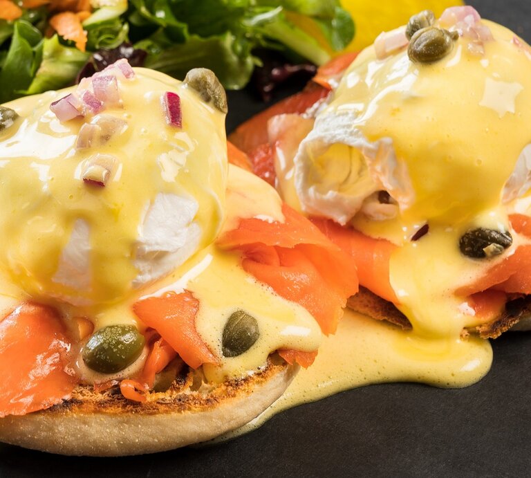 Die West-Coast-Eggs-Benedict auf schwarzem Untergrund mit einem kleinen Salat im Hintergrund. Alles seitlich und von leicht oben fotografiert. | © Shutterstock/ David Buzzard