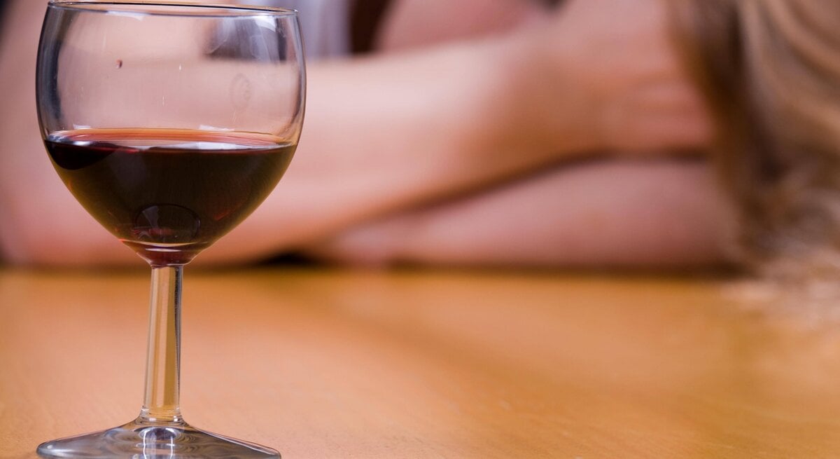 Ein Glas Rotwein auf dem Tisch, im Hintergrund eine verschwommene Person mit langen Haaren.