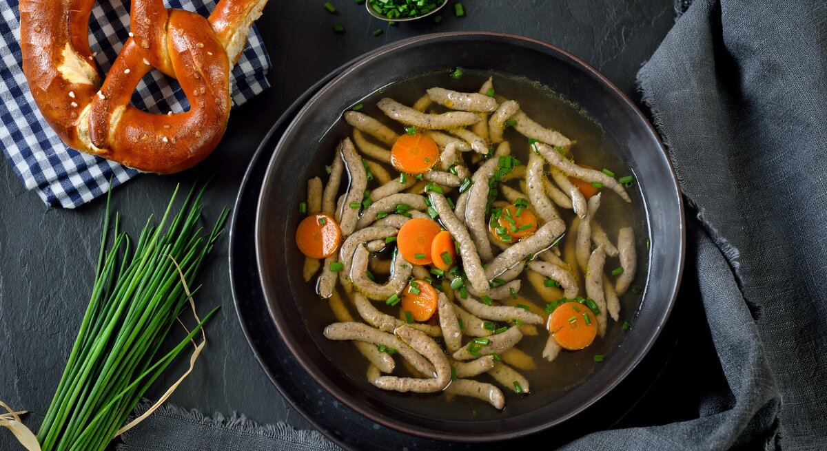 Draufsicht: Dunkler Untergrund, dunkles Tuch, daneben ein Teller bayerische Rindersuppe mit Leberspätzle. Daneben liegen Bund Schnittlauch, Schnittlauchröllchen und eine Breze auf einem kariertem Tuch.