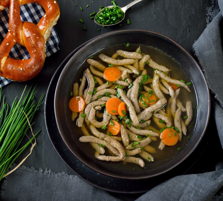 Draufsicht: Dunkler Untergrund, dunkles Tuch, daneben ein Teller bayerische Rindersuppe mit Leberspätzle. Daneben liegen Bund Schnittlauch, Schnittlauchröllchen und eine Breze auf einem kariertem Tuch. | © getty Images /  Bayerische Rindersuppe mit Leberspätzle