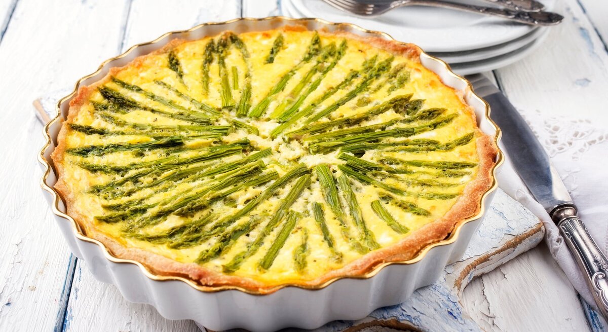 Eine vegane Spargelquiche in einer runden Quicheform auf weißem Holz, im Hintergrund Teller und Gabeln.