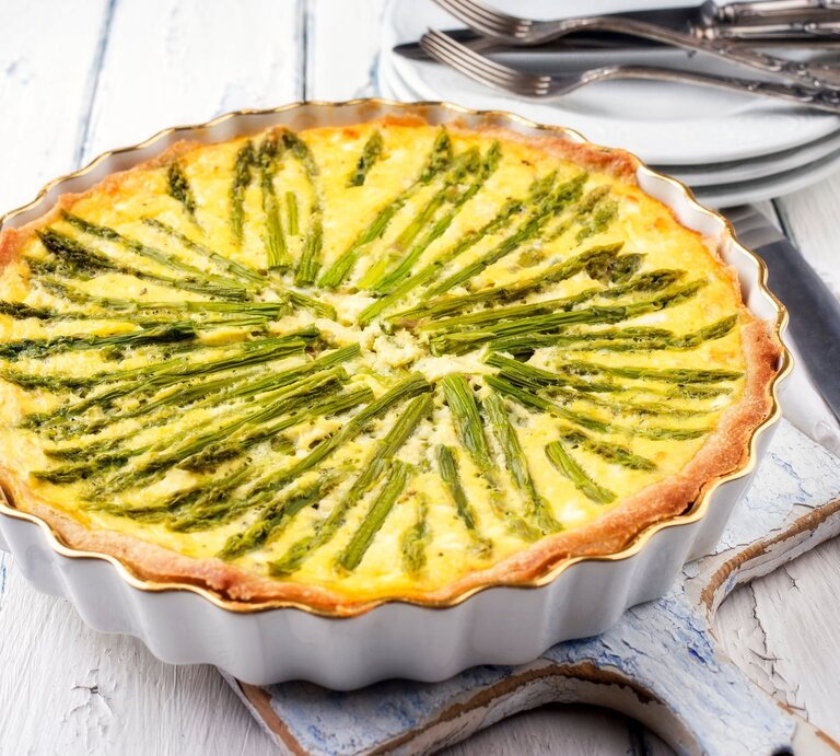 Eine vegane Spargelquiche in einer runden Quicheform auf weißem Holz, im Hintergrund Teller und Gabeln. | © Adobe Stock/ HLPhoto