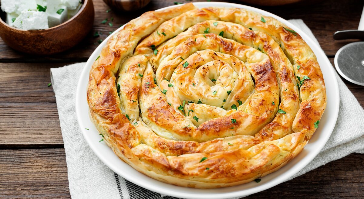 Die Spinat-Feta-Schnecke auf einem weißen Teller und dunklem Holztisch direkt von oben fotografiert. Daneben Feta und frische Kräuter.
