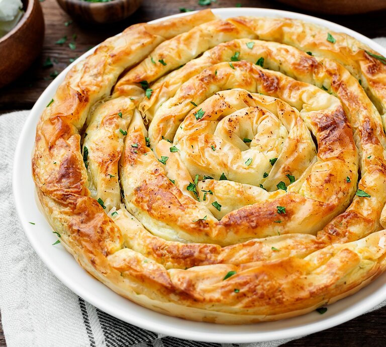 Die Spinat-Feta-Schnecke auf einem weißen Teller und dunklem Holztisch direkt von oben fotografiert. Daneben Feta und frische Kräuter. | © Shutterstock/ nelea33 