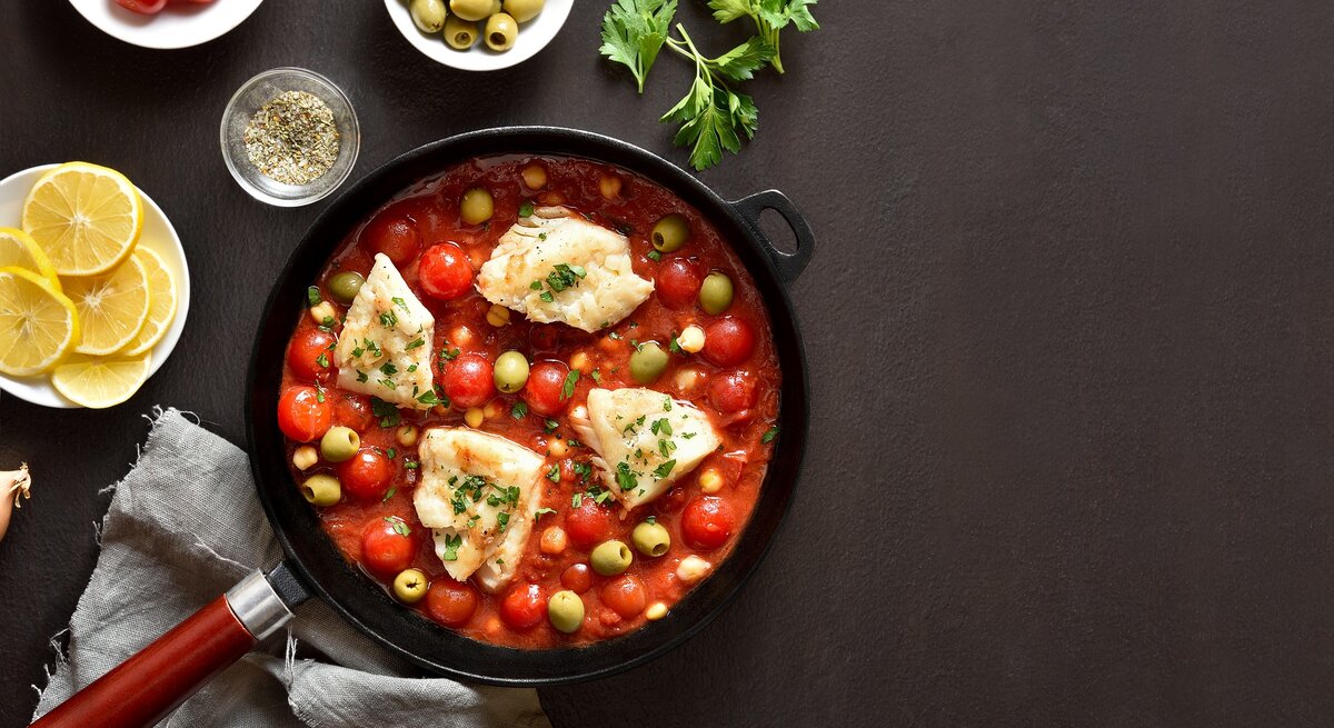 Draufsicht: Eine mediterrane Fischfilet-Pfanne auf dunklem Untergrund. Drum herum liegen Zitronenscheiben, Petersilie und Gewürze.
