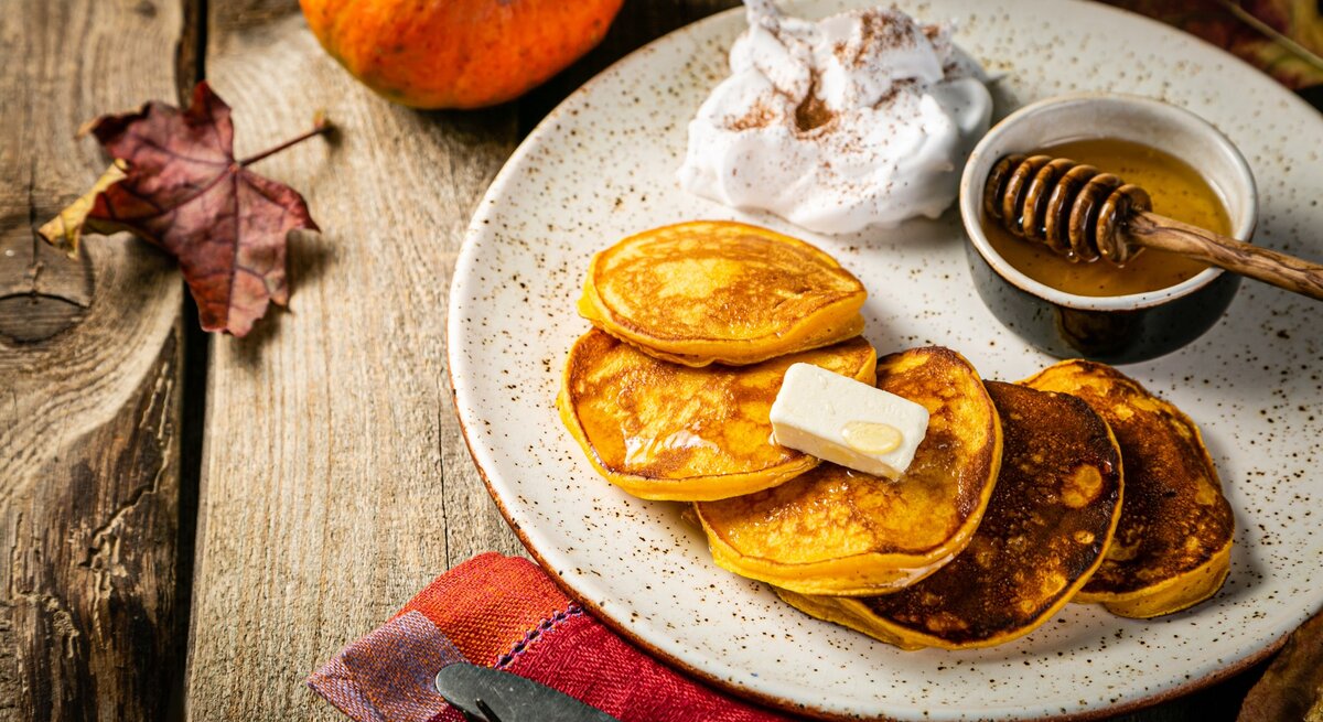Vier Kürbis Pancakes auf einem hellen Teller mit Honig und etwas geschlagener Sahne. Alles von schräg oben auf einem rustikalen Holztisch mit herbstlicher Deko fotografiert.